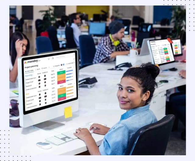 A smiling woman at her desk beside a monitor showing a monday.com project planning board