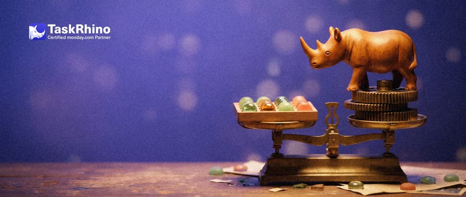 A small wooden rhino weighs a heavy block of brass gears against a tidy board of coloured tiles on an old balance scale.
