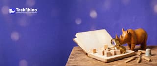 A small wooden rhino studies a large open blank notebook with small wooden building blocks stacked on its pages, holding a brass loupe.