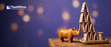 A small wooden rhino props up a six-level house of cards that is leaning and about to topple with a small wooden stick.