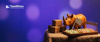 A small wooden rhino holds a tiny chequered flag at a finish line between a stack of many small matching boxes and a tidy board of coloured tiles.
